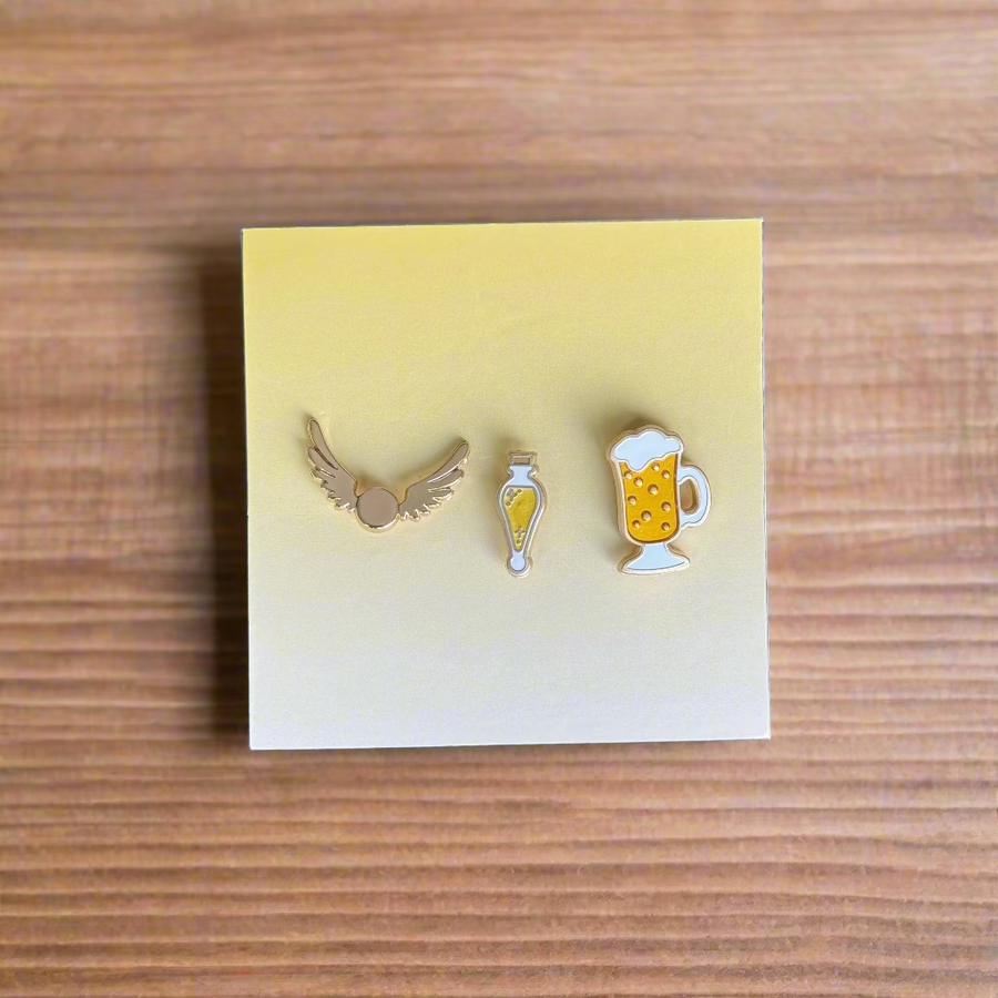 Set of three mini enamel pins: golden winged ball, lemon potion vial, and frothy butter-brew mug on display card.