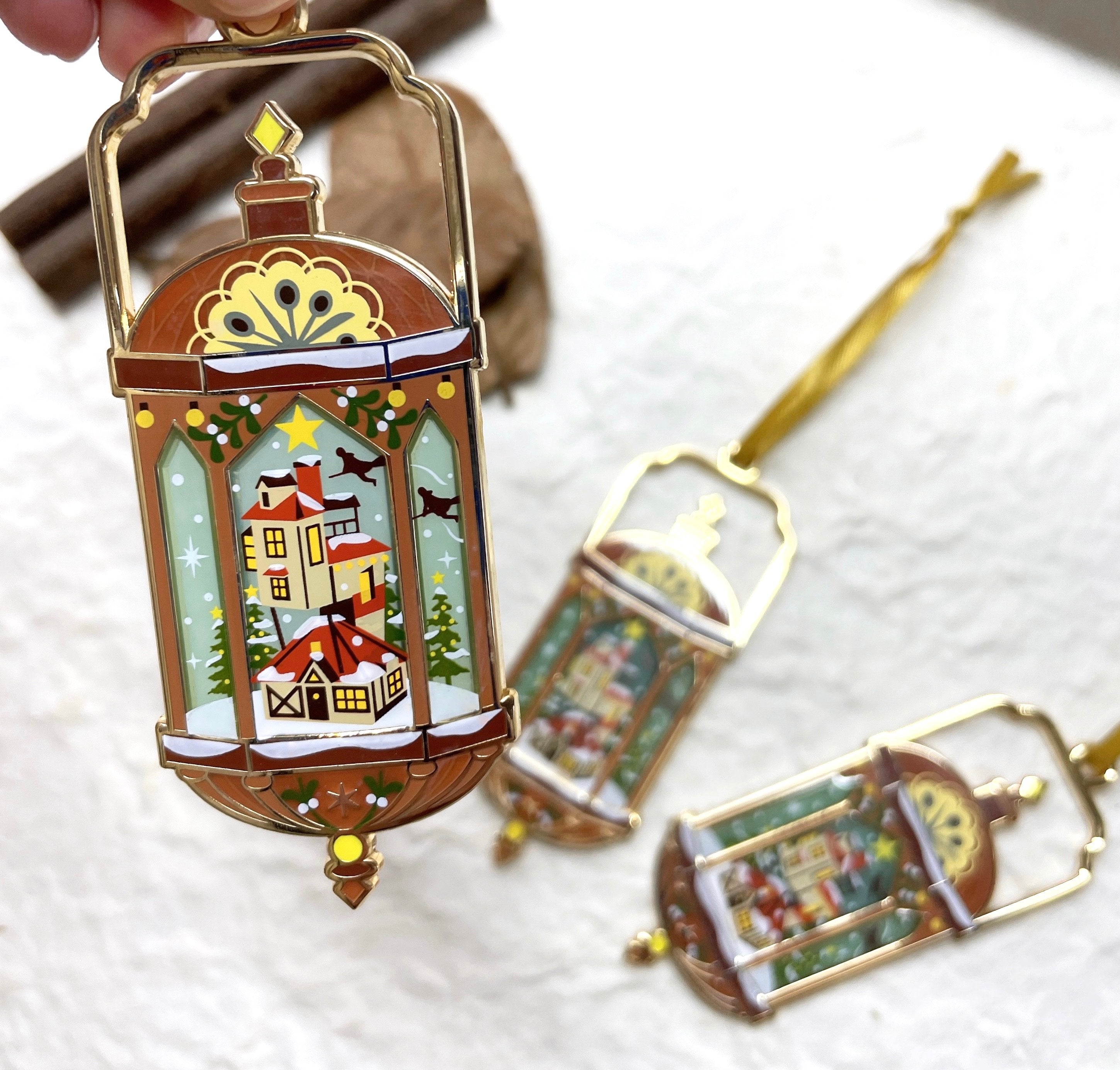 Lantern ornament holding a snow‑covered cottage scene with pine trees—hanging loop and antique black frame.